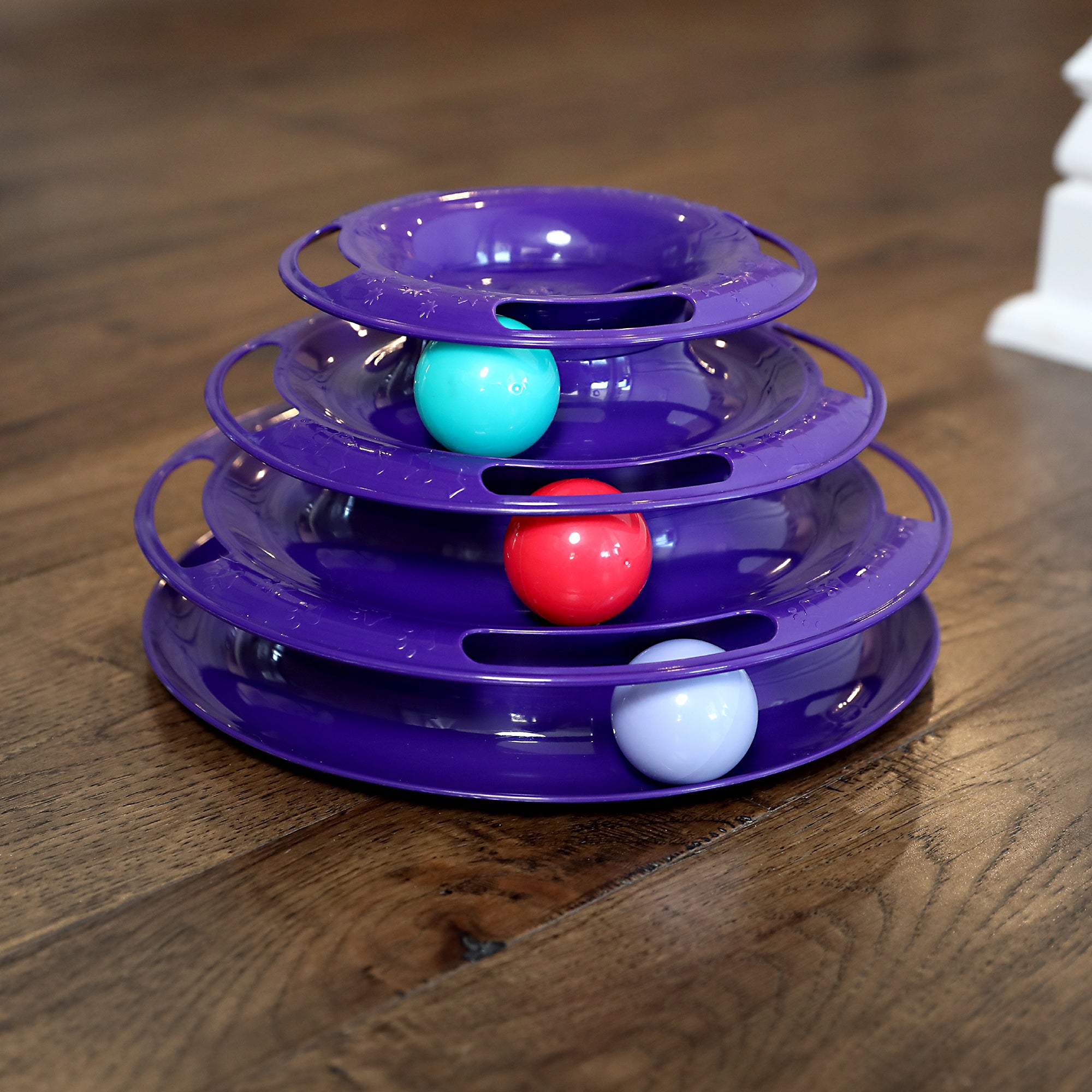 The Quirky Kitty Three Layer Ball Track Cat Toy by Quirky Kitty is a purple, tiered plaything with three levels featuring vibrant turquoise, red, and white balls. It offers engaging interactive play while resting on a polished wooden floor.