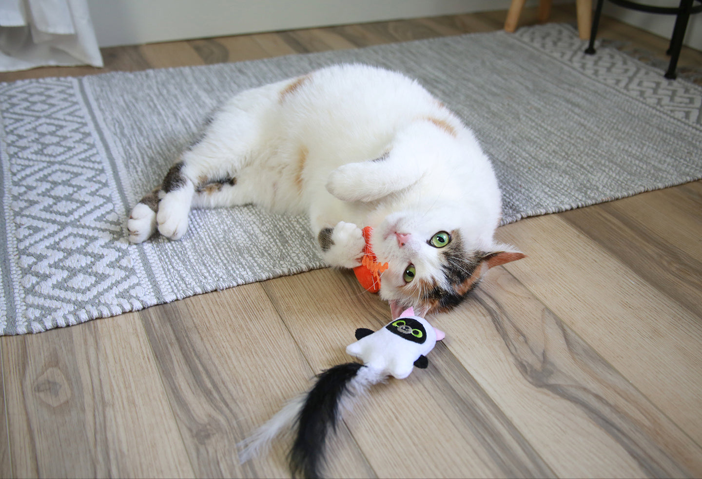 A fluffy white cat with brown patches playfully rolls on a gray patterned rug, clutching the Mad Cat Hiss & Haunt Plush Cat Toy in its paws. Nearby, a Halloween wand with a feathered tail enticingly lies on the wooden floor.