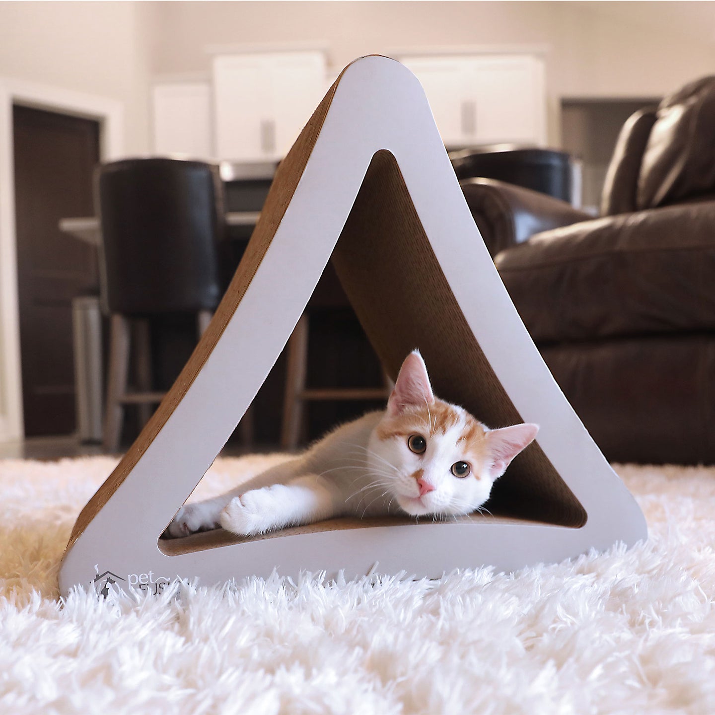 A white and orange cat playfully lies inside a triangular PetFusion 3-Sided Vertical Cat Scratching Post on a fluffy white rug. The cozy living room in the background features a dark leather chair and visible kitchen, highlighting stylish PetFusion pet furniture made from recycled cardboard.