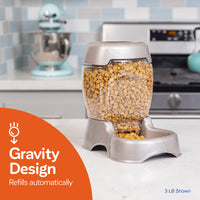 A Petmate Pet Cafe Feeder with a clear hopper filled with dry kibble sits on the kitchen counter, ensuring automatic refills. Behind it are a mixer and stove, accompanied by an orange graphic stating Gravity Design Refills Automatically.