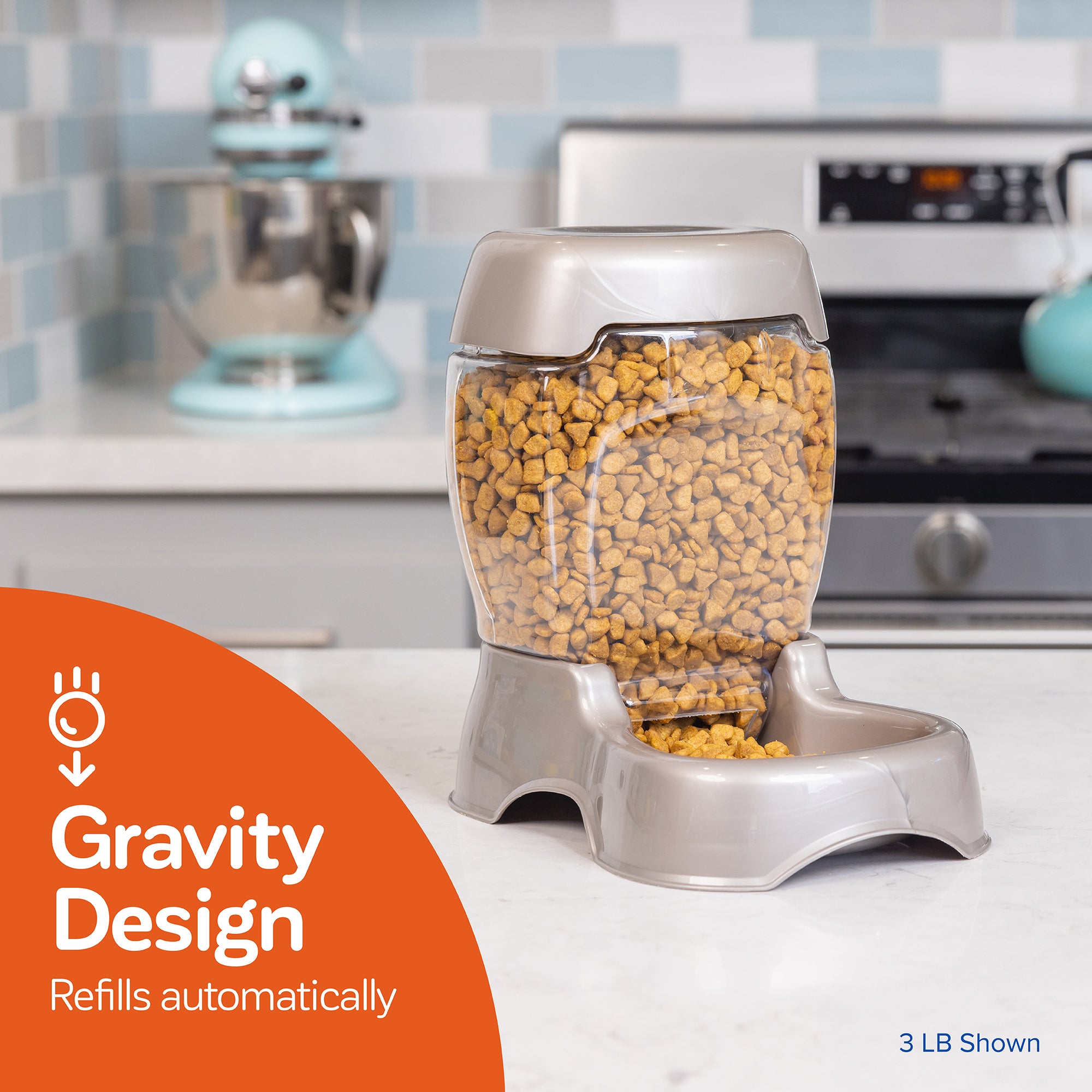 A Petmate Pet Cafe Feeder with a clear hopper filled with dry kibble sits on the kitchen counter, ensuring automatic refills. Behind it are a mixer and stove, accompanied by an orange graphic stating Gravity Design Refills Automatically.