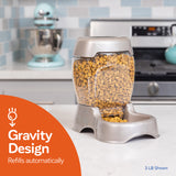 A Petmate Pet Cafe Feeder with a clear hopper filled with dry kibble sits on the kitchen counter, ensuring automatic refills. Behind it are a mixer and stove, accompanied by an orange graphic stating Gravity Design Refills Automatically.