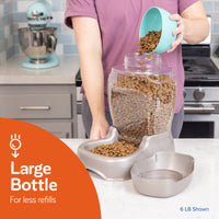 A person pours pet food from a teal bowl into the Petmate Pet Cafe Feeder, which is a large transparent gravity feeder with an attached gray dish for reduced refill frequency, sitting on the kitchen counter. In the background, theres a mixer and a stove. 6 LB Shown.