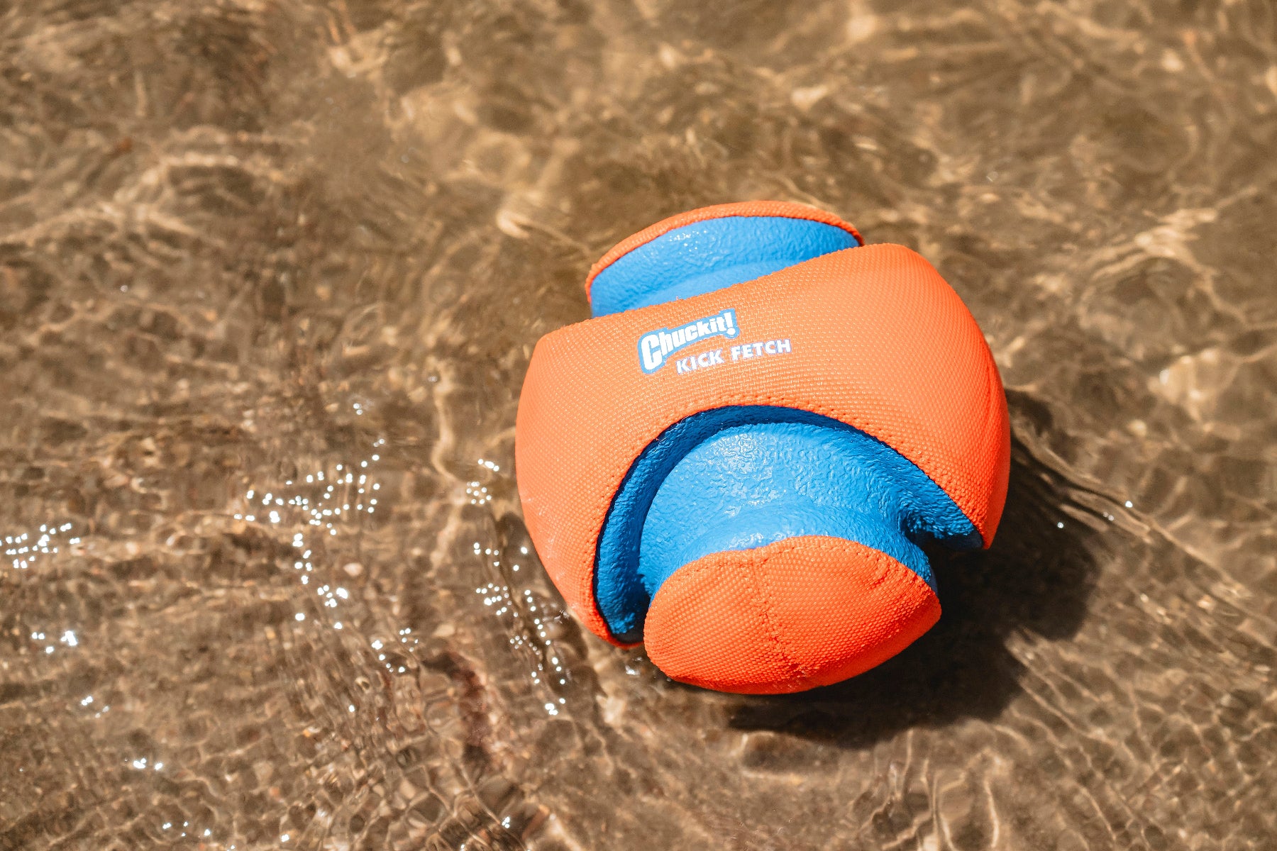 A Chuckit Kick Fetch Dog Toy, with its vibrant orange and blue design, floats in clear, shallow water. Surrounded by ripples that highlight its texture and colors, it is perfect for interactive fun with your dog.