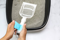 An individual uses the Arm & Hammer Deluxe 2-In-1 Litter Scoop with Waste Bags over a black litter box filled with gray, sand-like litter. In the background, against a white surface, Arm And Hammer baking soda showcases its odor-neutralizing power.