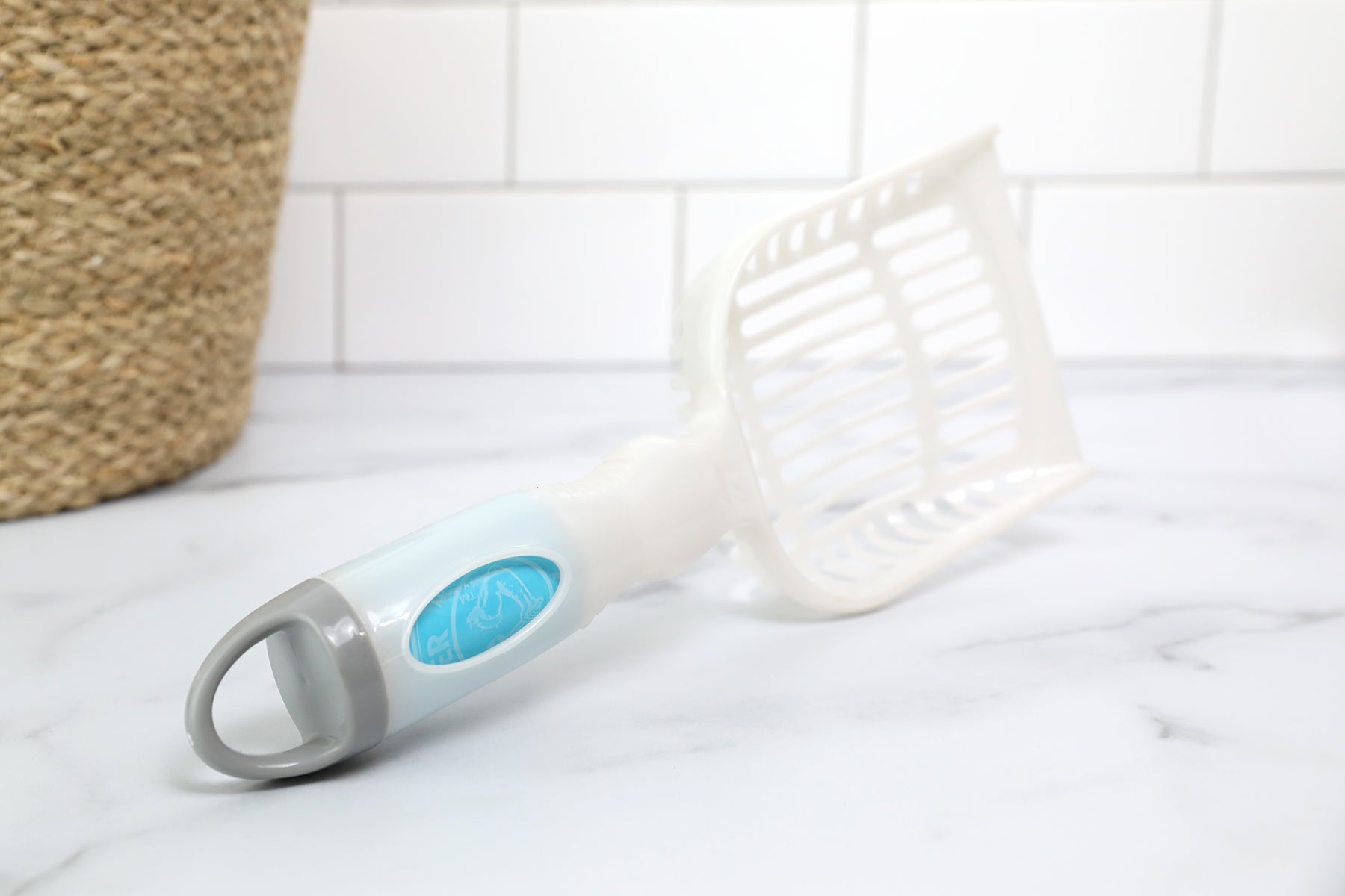 An Arm & Hammer Deluxe 2-In-1 Litter Scoop with Waste Bags sits on a marble countertop, showcasing its white and light blue plastic build, slotted design, and curved handle. A woven basket against the tiled wall is ideal for storing its odor-neutralizing bags to maintain freshness.