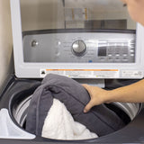 A gray and white blanket is placed into a top-loading washing machine to clean the PetFusion Calming Cuddler Dog Bed. The control panel settings are visible through the glass lid, offering a reassuring view inside while preparing this anxiety-relief space for dogs.