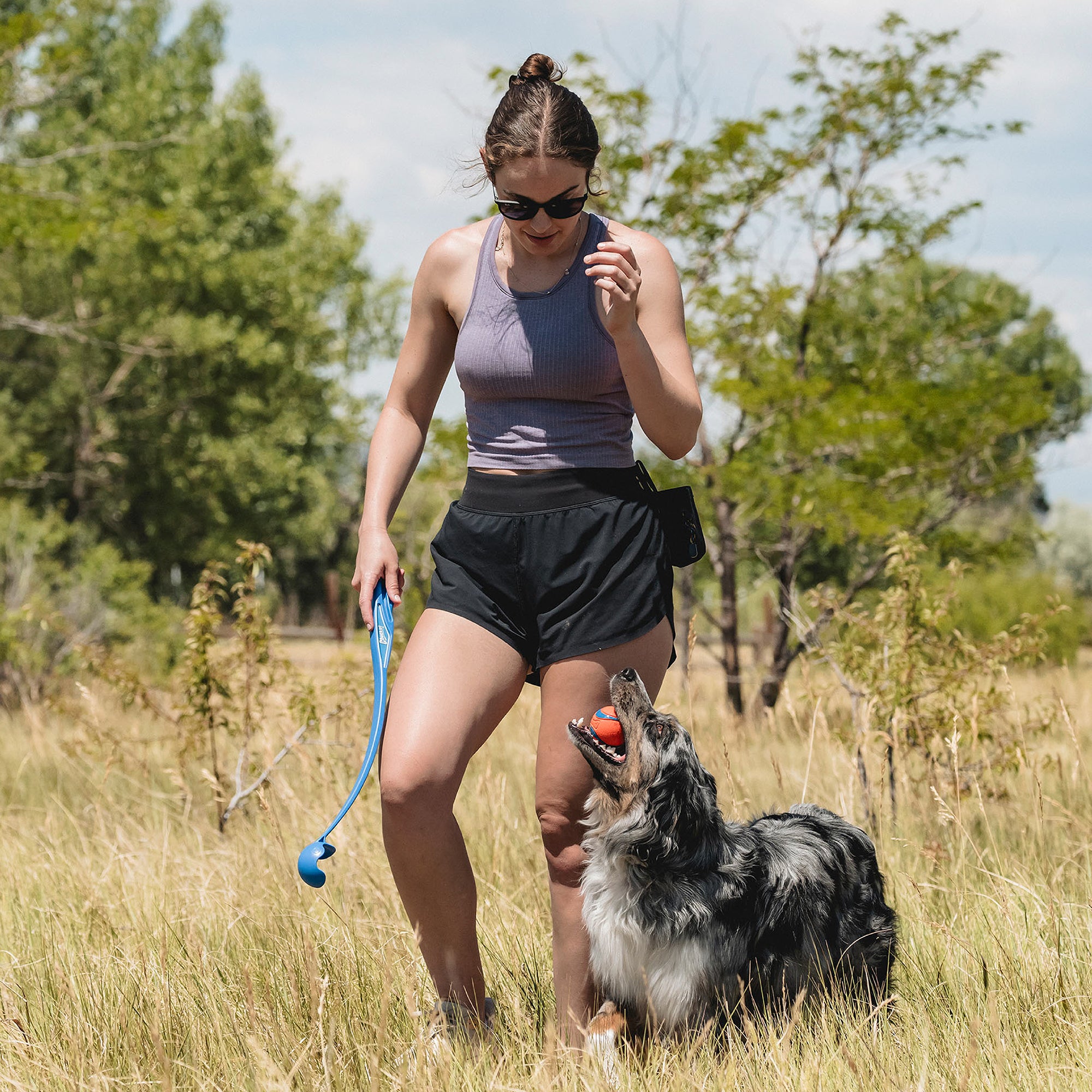 A woman in sunglasses and athletic wear stands in a grassy field, holding the Petmate Chuckit! Sport 18M Launcher with Ultra Ball Medium 3 Pack, as her dog with a ball looks up at her amid surrounding trees and greenery.