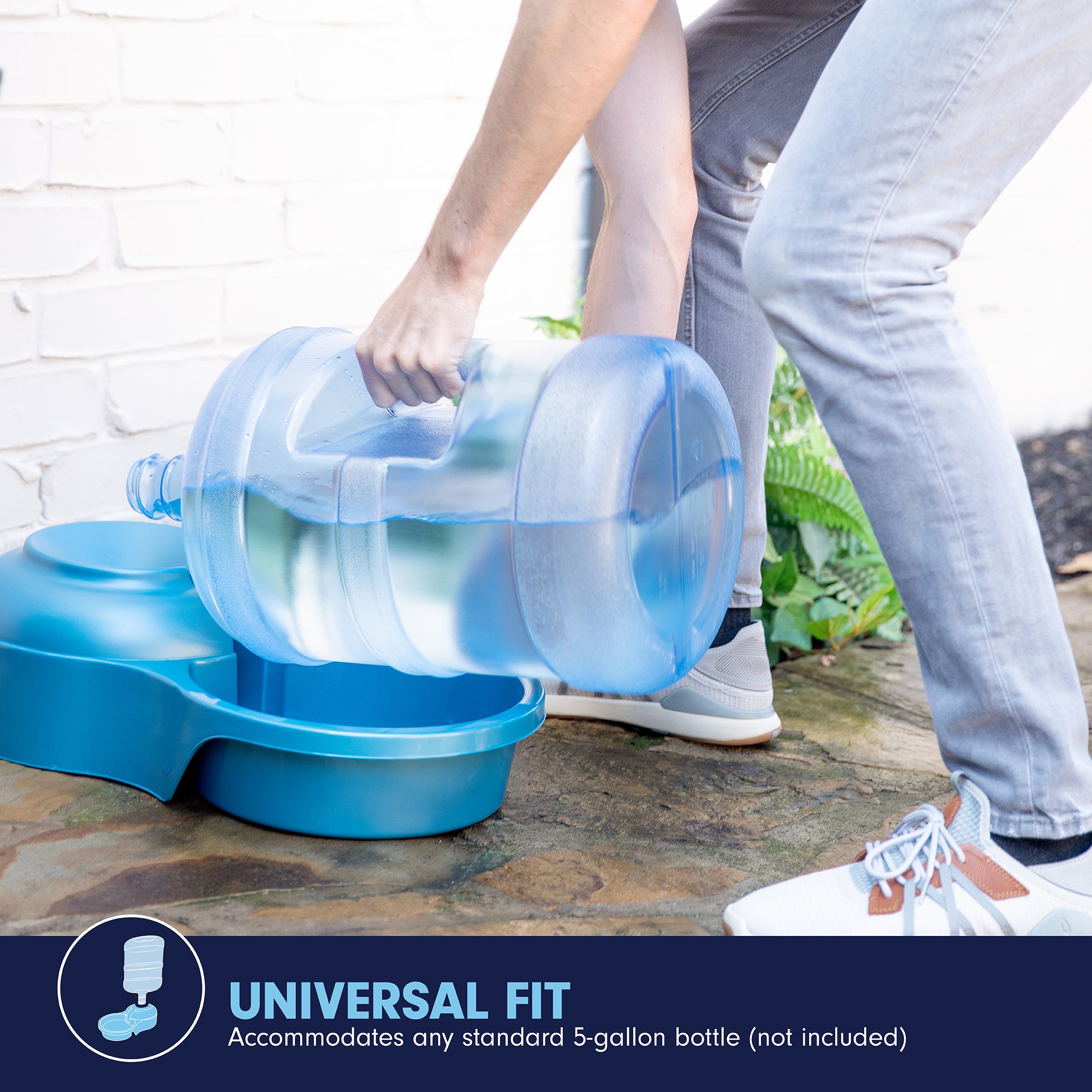 Wearing jeans and sneakers, a person places a large clear 5-gallon water bottle into the Petmate Big Blue Bowl, a blue pet water dispenser. Perfect for outdoor hydration, it accommodates any standard 5-gallon bottle (not included), ensuring pets always have fresh water.