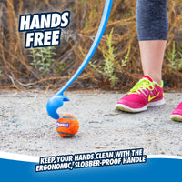 Wearing pink sneakers, someone uses a blue Chuckit! Junior 18M Dog Ball Launcher to pick up an orange ball on a gravel path. With Hands Free text highlighting its ergonomic, slobber-proof handle, it keeps hands clean against a backdrop of grass and plants.