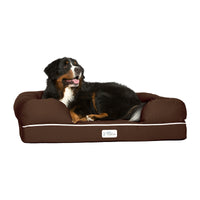 A large black, brown, and white dog relaxes on a PetFusion Ultimate Dog Bed With Memory Foam in brown. The bed has raised cushioned edges for joint pain relief. The relaxed dog lies on its side with its mouth slightly open, while a small label with the Petfusion logo is visible on the bed.
