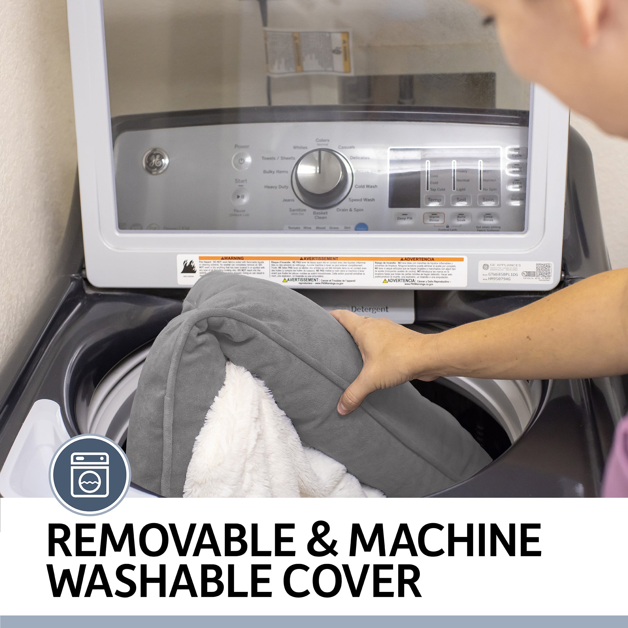 A person puts a gray and white cushion cover from the PetFusion Ultimate Dog Bed With Memory Foam into a top-loading washer. The control panel is visible with text reading: Removable & Machine Washable Cover, ensuring easy maintenance for anxiety support and joint pain relief.