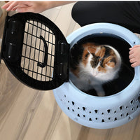A calico cat relaxes inside the Petmate Get-A-Round E-Z Load Kennel For Cats, enjoying a secure, comfortable spot as a person kneels nearby on the light wooden floor.
