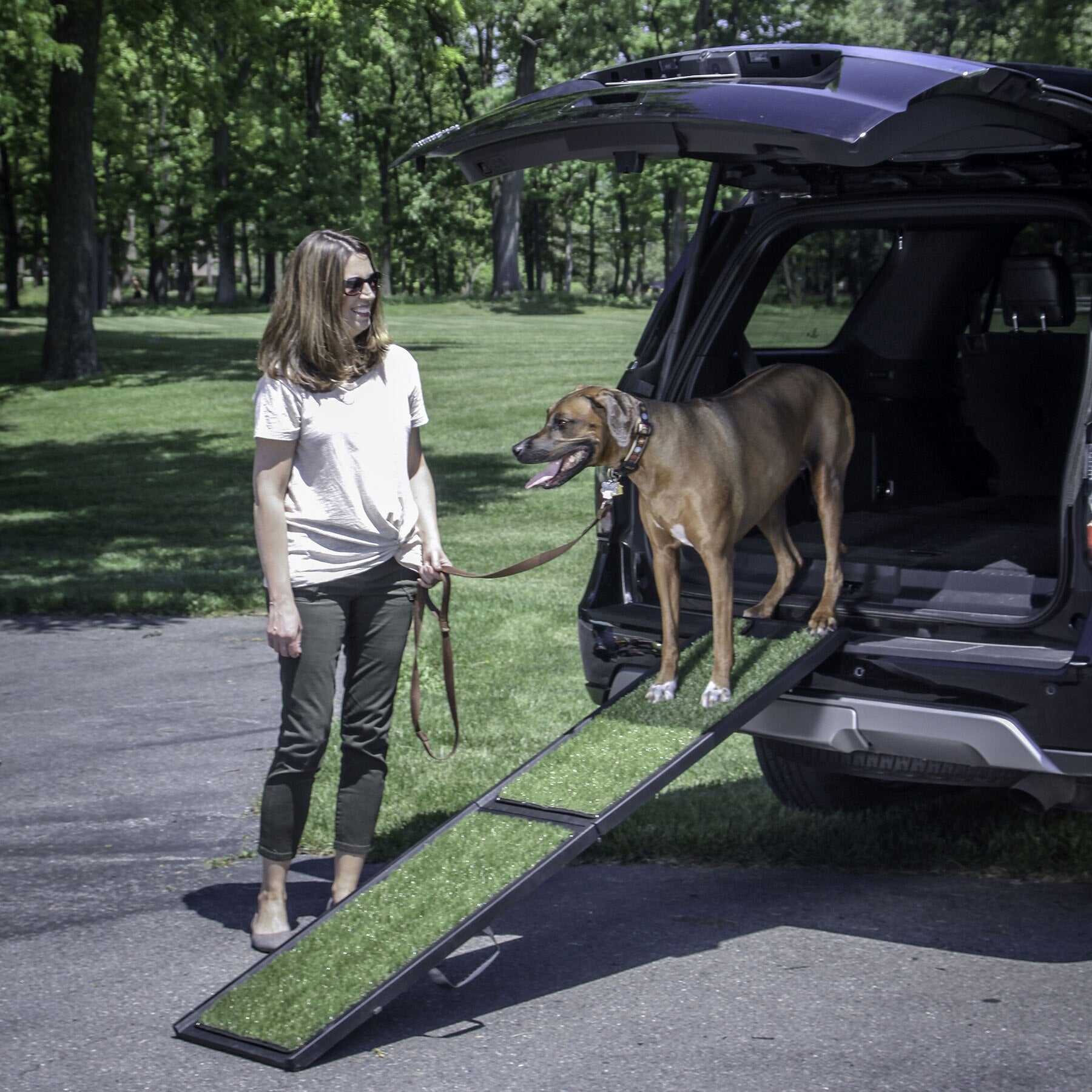 Beside an SUV at a sunny, tree-lined park, a woman smiles as her large brown dog happily trots down the Gen7 Gen7Pets Natural Step Ramp for Vehicles, from the open trunk onto the green grass.