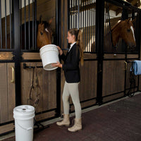 In a stable, a woman in riding attire stands near a brown horse with the Vittles Vault Outback Pet Food Storage Bucket. Another horse is in the adjacent stall surrounded by gear, with a blue towel nearby. The scenario suggests attentive care supported by secure food storage solutions from Vittles Vault.