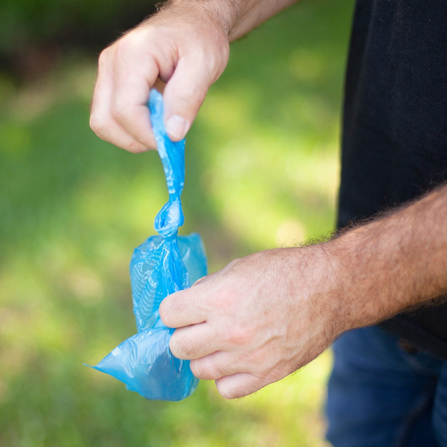 Someone expertly ties a knot in a small, blue Arm & Hammer Easy-Tie Dog Waste Bag outdoors. The grassy background is blurred, highlighting the hands and the bag designed for optimal odor control by Arm And Hammer.