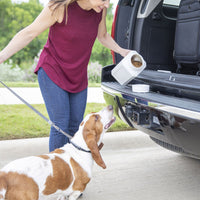 A woman in a maroon sleeveless top and jeans feeds her brown and white Basset Hound from a BPA-free Vittles Vault Junior by Vittles Vault at the open back of her vehicle. The dog is on a leash, enjoying his meal with a grassy backdrop.