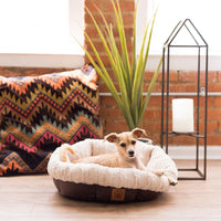 A small tan dog lies comfortably in a SnooZZy Rustic Luxury Ultra Cuddler Pet Bed, featuring a plush, round design and non-skid bottom. The setting includes a colorful cushion, tall green plant, and candle in a metal holder against a softly lit brick wall.