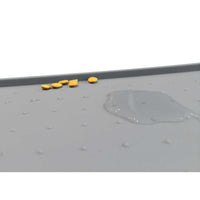 A gray PetFusion Pet Food Mat made from FDA-grade premium silicone with anti-slip bumps displays scattered yellow pills on the left and a puddle of clear liquid on the right.