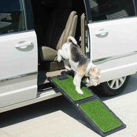 A small dog descends from a white minivan using the Gen7Pets Natural Step Ramp for Vehicles, featuring a poly-grass surface. The car door is open, and the scene is set under the sunny outdoors.